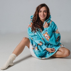 SNUGLFIT™ Hoodie Blanket - Sporty Corgi design, cozy oversized wearable blanket, modeled by a smiling woman sitting cross-legged indoors.